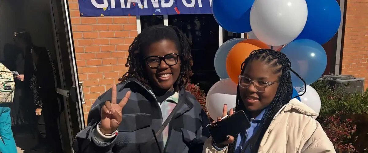 Two students holding plates of food, holding up peace signs with fingers and smiling outside of brick building