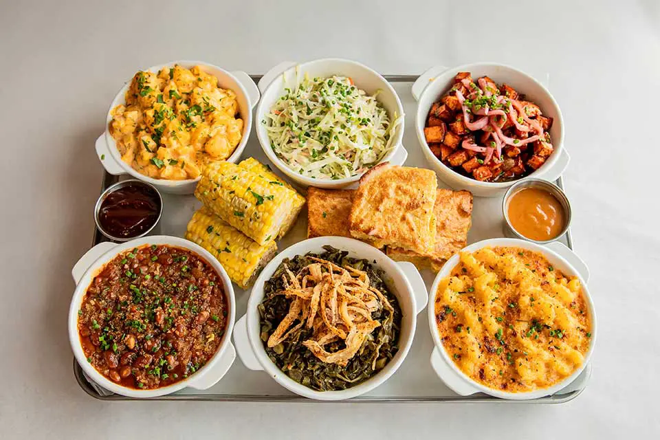 A tray with seven bowls of various Southern-style side dishes, including macaroni and cheese, coleslaw, baked beans, candied yams, collard greens topped with fried onions, cornbread, and barbecue sauce, all arranged neatly on a light-colored surface.